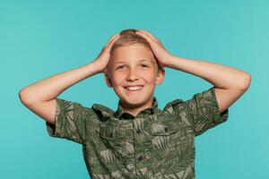 Close-up portrait of a blonde teenage boy in a green shirt with palm print posing against a blue studio background. Concept of sincere emotions. photo