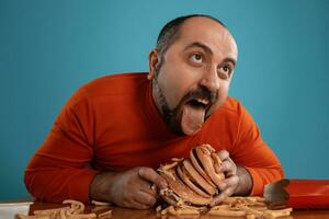 Close-up portrait of a middle-aged man with beard, dressed in a red turtleneck, posing with burgers and french fries. Blue background. Fast food. photo