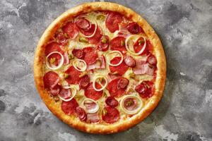 Pizza with bacon slices, salami, hunting sausages, jalapeno and onions on gray background photo