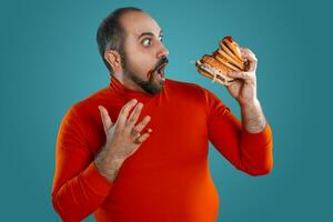 Close-up portrait of a middle-aged man with beard, dressed in a red turtleneck, posing with burgers against a blue background. Fast food. photo