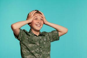 Close-up portrait of a blonde teenage boy in a green shirt with palm print posing against a blue studio background. Concept of sincere emotions. photo