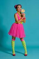 Lovely girl with a multi-colored braids hairstyle and bright make-up, posing in studio against a blue background, holding a lollipop in her hand. photo