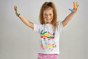 Little girl in white painted t-shirt is posing standing isolated on white and gesticulating with her colored in different paints palms and face. Art studio. Close-up. photo