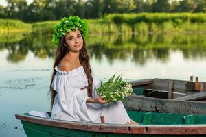 The nymph with long dark hair in a white vintage dress sitting in a boat in the middle of the river. photo