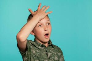 Close-up portrait of a blonde teenage boy in a green shirt with palm print posing against a blue studio background. Concept of sincere emotions. photo