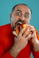 Close-up portrait of a middle-aged man with beard, dressed in a red turtleneck, posing with burgers against a blue background. Fast food. photo