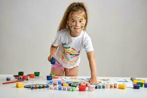 Little girl in white t-shirt standing at table with whatman and colorful paints, painting on it with her hands. Isolated on white. Medium close-up. photo