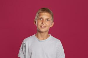 Close-up portrait of a blonde teenage boy in a white t-shirt posing against a pink studio background. Concept of sincere emotions. photo