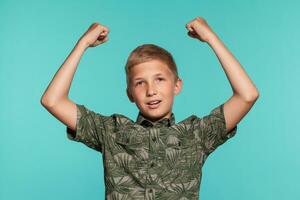 de cerca retrato de un rubia Adolescente chico en un verde camisa con palma impresión posando en contra un azul estudio antecedentes. concepto de sincero emociones foto