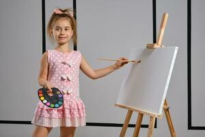 Little girl artist in a pink dress is standing behind easel and painting with brush on canvas at art studio with white walls. Medium close-up shot. photo