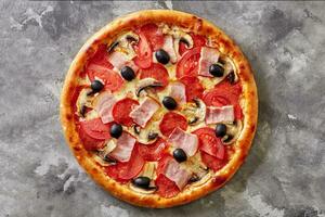 Thin pizza with bacon, salami, mushrooms, tomatoes and black olives on gray stone surface photo