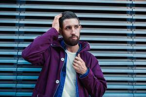 Handsome young man with hairstyle in fashionable clothes stands near iron wall and looking sideways photo