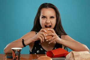 de cerca retrato de un mujer en un negro vestir posando sentado a el mesa con hamburguesas, francés papas fritas y beber. azul antecedentes. rápido alimento. foto