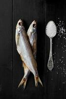 Two air-dried sabrefish on black wooden surface with spoonful of salt photo