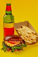 Cheeseburger with beef patty, tomatoes, onion and greens with French fries and bottle drink isolated on yellow background photo