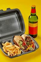 Takeaway container with vegan burrito and fries on yellow background with bottle of drink photo