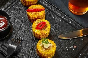 Slices of cooked sweetcorn on cob with salt, butter, chili peppers and ketchup on slate board photo