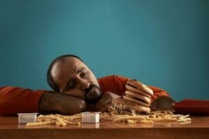 de cerca retrato de un de edad mediana hombre con barba, vestido en un rojo suéter tipo con cuello de tortuga, posando con hamburguesas y francés papas fritas azul antecedentes. rápido alimento. foto