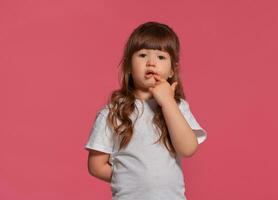 de cerca retrato de un pequeño morena niña vestido en un blanco camiseta posando en contra un rosado estudio antecedentes. sincero emociones concepto. foto