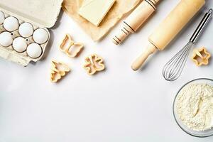Bowl with wheat flour, rolling pin, whisk, eggs, butter, cookie cutters. Top view on a white table with a copy space photo