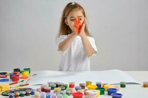 Little girl in white t-shirt sitting at table with whatman and colorful paints on it, painting her hands. Isolated on white. Medium close-up. photo