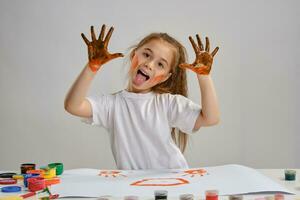 Little girl in white t-shirt sitting at table with whatman and colorful paints, showing her painted hands. Isolated on white. Medium close-up. photo