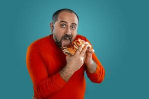 Close-up portrait of a middle-aged man with beard, dressed in a red turtleneck, posing with burgers against a blue background. Fast food. photo