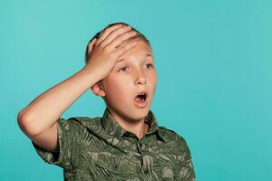 Close-up portrait of a blonde teenage boy in a green shirt with palm print posing against a blue studio background. Concept of sincere emotions. photo