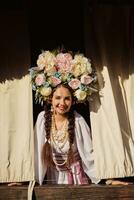 Brunette girl in a white ukrainian authentic national costume and a wreath of flowers is is looking out of the window. Close-up. photo