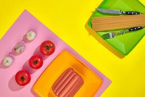 A lot of raw sausages on plate. On yellow background with pasta and vegetables, top view. photo