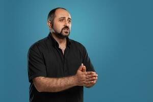 Close-up portrait of a brunet middle-aged man with beard, dressed in a black t-shirt and posing against a blue background. photo