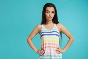 Brunette woman with long hair, dressed in colorful striped shirt, posing against blue studio background. Sincere emotions. Close-up. photo