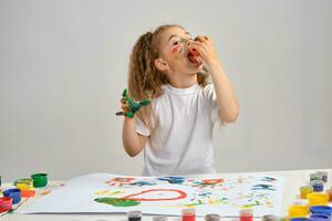 Little girl in white t-shirt sitting at table with whatman and paints on it, posing with painted face and hands. Isolated on white. Medium close-up. photo