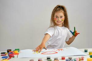 Little girl in white t-shirt sitting at table with whatman and colorful paints, painting on it with her hands. Isolated on white. Medium close-up. photo