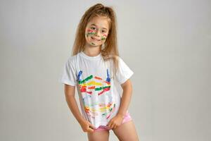 Little girl in white painted t-shirt is posing standing isolated on white and gesticulating with her colored in different paints palms and face. Art studio. Close-up. photo