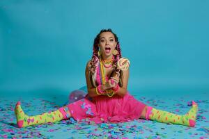 encantador niña con un multicolor trenzas peinado y brillante constituir, posando en estudio con chupete, aire globos y papel picado en contra un azul antecedentes. foto