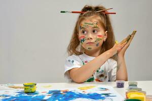 Little girl in white t-shirt, with brushes in her hair is sitting at the table with whatman and paints, painting on it. Isolated on white. Close-up. photo