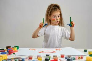 Little girl in white t-shirt sitting at table with whatman and colorful paints, painting on it with hands, face. Isolated on white. Medium close-up. photo