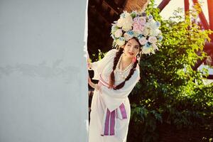 Brunette girl in a white ukrainian authentic national costume and a wreath of flowers is posing against a white hut. photo