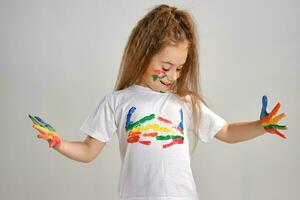 Little girl in white painted t-shirt is posing standing isolated on white and gesticulating with her colored in different paints palms and face. Art studio. Close-up. photo