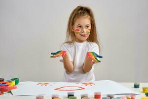 Little girl in white t-shirt sitting at table with whatman and colorful paints, showing her painted hands, face. Isolated on white. Medium close-up. photo