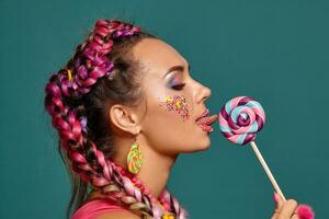 encantador niña con un multicolor trenzas peinado y brillante constituir, posando en estudio en contra un azul fondo, participación un pirulí en su mano. foto