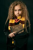 Portrait of a little witch girl with long brown hair dressed in dark coat, holding magic wand in her hand, posing on black studio background. photo