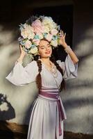 morena niña en un blanco ucranio auténtico nacional disfraz y un guirnalda de flores es posando en contra un blanco cabaña. de cerca. foto