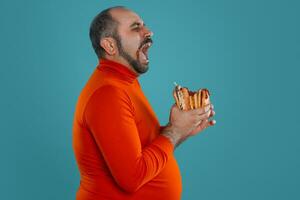 Close-up portrait of a middle-aged man with beard, dressed in a red turtleneck, posing with burgers against a blue background. Fast food. photo