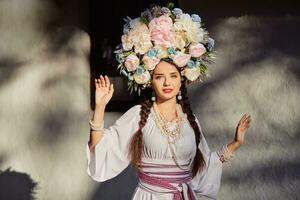 morena niña en un blanco ucranio auténtico nacional disfraz y un guirnalda de flores es posando en contra un blanco cabaña. de cerca. foto