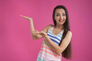 Brunette woman with long hair, dressed in colorful striped shirt, posing against pink studio background. Sincere emotions. Close-up. photo