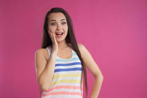 Brunette woman with long hair, dressed in colorful striped shirt, posing against pink studio background. Sincere emotions. Close-up. photo