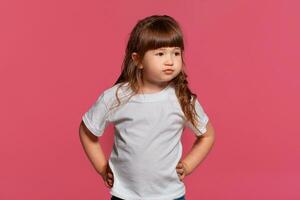 de cerca retrato de un pequeño morena niña vestido en un blanco camiseta posando en contra un rosado estudio antecedentes. sincero emociones concepto. foto