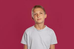 Close-up portrait of a blonde teenage boy in a white t-shirt posing against a pink studio background. Concept of sincere emotions. photo